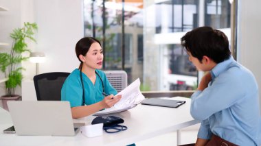 Önlüklü Asyalı kadın doktor klinikteki genç Asyalı erkek hastalara sağlık raporunu açıklıyor. Doktor tavsiyeler için teşhis tedavisi ve sağlık hizmetleri hakkında danışmanlık yapar.