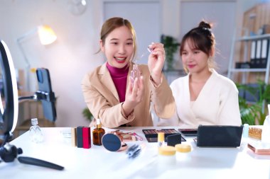 İki genç Asyalı kadın makyaj ve cilt bakımı için canlı yayın yapan güzellik ipuçları sunuyor. Halka ışığı ve akıllı telefonla çevrili beyaz bir masada ürünleri gösteriyorlar.