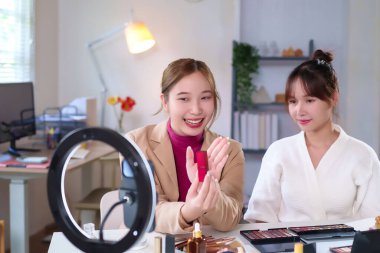 İki genç Asyalı kadın, sosyal medya kanallarında ruj incelemesi yapan bir makyaj dersini canlı yayınlıyorlar. Biri ruj tutuyor, diğeri de izliyor. Güzellik ve kozmetik ürünlerini tanıtıyorlar..