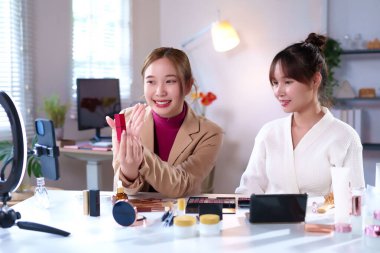 İki Asyalı yetişkin kadın makyaj dersinde canlı yayında. Biri ruj sergilerken diğeri cilt bakımını tartışıyor. Sosyal medya için güzellik blogcuları olarak içerik yaratıyorlar, yeni güzellik ürünlerini gözden geçiriyorlar ve makyaj malzemelerini gösteriyorlar. 