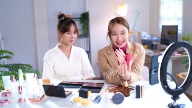 İki genç Asyalı kadın sosyal medya için makyaj eğitimi ve ürün değerlendirmesi yapıyor. Takipçileriyle güzellik ipuçlarını paylaşıyorlar. Bir kadın elinde ruj tutuyor..