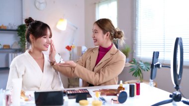 İki Asyalı kadının yakalanması, biri diğerine parlak bir iç mekanda makyaj yapıyor güzellik tavsiyelerini paylaşıyor ve ışık yaymak için kullanılan bir halkayla içerik oluşturuyor.