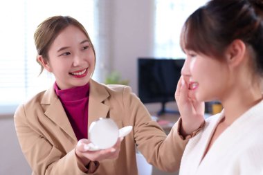 Modern bir ofiste ya da ev ortamında iki genç Asyalı kadın. Biri, diğerine güzellik tedavisiyle ilgili kozmetik bir ürün sunuyor, sağlıklı bir yaşam tarzı ve sağlığını destekliyor..