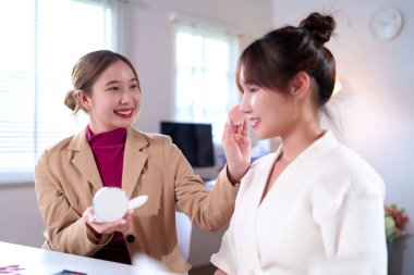 Bu resim genç Asyalı kadınları güzellik rutininde gösteriyor. Biri diğerine makyaj yapıyor, kozmetik uygulama ve cilt bakımını gösteriyor. Evdeki kozmetoloji hizmetini vurguluyor