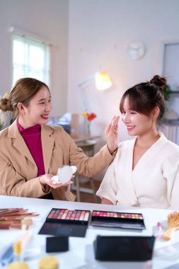 Evde ya da güzellik salonunda iki Asyalı kadın. Genç makyaj sanatçısı, çeşitli kozmetik ürünleri sergilenerek mükemmel görünüm elde etmek için sünger kullanarak modeline temel oluşturur..