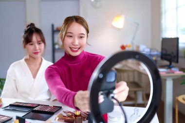 İki Asyalı kadın, sosyal medya kanalları için halka ışığıyla makyaj dersleri hazırlıyorlar. Yeni başlayanlar ve profesyoneller için makyaj malzemesi değerlendirmeleri gösteriliyor.