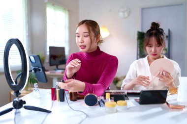 İki genç Asyalı kadın güzellik blogu için makyaj eğitimi hazırlıyorlar. Masada yüzük ışığı ve cep telefonu var. Çevrimiçi etkili canlı yayında. Sosyal medya için çevrimiçi içerik oluştur.