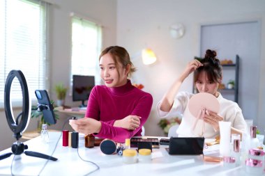 İki genç Asyalı kadın ev stüdyosunda kozmetik ve halka ışığıyla güzellik içeriği yaratıyor. Sosyal medya platformu için çevrimiçi makyaj dersi canlı yayın videosu.
