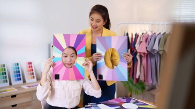 İki Asyalı kadın kıyafet seçimleri için kişisel renk paletlerini inceliyor. Bir kadın, sezonluk renk analizi eğilimleri için yüzünün yanında bir palet çerçeve tutuyor..