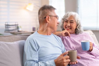 Yaşlı bir Asyalı çiftin evde sakin bir anın tadını çıkarırken çekilmiş hoş bir fotoğrafı. Gülümsüyorlar, kahve içiyorlar ve emeklilik yıllarını birlikte geçiriyorlar..