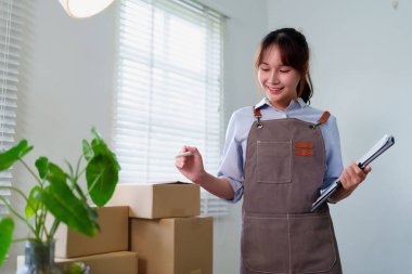 Bu, önlük takan Asyalı genç bir girişimcinin karton kutuları online mağaza işi için hazırlarken ki görüntüsü. O, ev ofisinde çalışıyor..