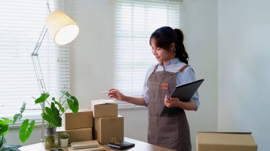 Genç Asyalı kadın paketleri müşterilere göndermek için hazırlıyor. El yapımı ürünler satan bir online mağazası var. Paketleri hazırlamak için siparişleri kontrol ediyor..