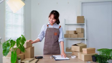 Önlüklü gülümseyen Asyalı genç bir kadın, çevrimiçi dükkan müşterilerine kaliteli hizmet sunarak ev ofisinden nakliye için paketler hazırlıyor..