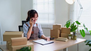 Genç Asyalı iş kadını ev ofisinde telefonla emirler alıyor. Çevrimiçi satış, ekommerce ve karton nakliye kutularıyla teslimatı yönet.