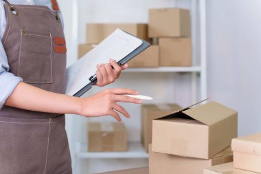 Genç bir Asyalı girişimci teslimat için paketler hazırlıyor. KOBİ ya da küçük işletmeler için ev deposundaki kalemle listeyi kontrol ediyor.