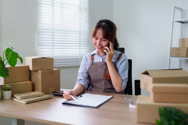 Asyalı genç bir kadın telefonuyla bir müşteriyle konuşmaktan mutlu. Ev ofisindeki hisseleri kontrol ediyor. Girişimcisiyle online bir iş yürütüyor..