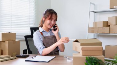 Genç Asyalı girişimci kadın evdeki ofisinde kutu paketlerken telefon kullanıyor. Çevrimiçi pazarlama ve sipariş teslimatı ile küçük işletme girişimi yönetiliyor.