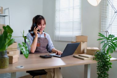 Akıllı telefondan konuşan genç Asyalı bir kadın çevrimiçi iş için dizüstü bilgisayarda çalışırken ve evden gelen siparişleri yönetirken küçük işletmeleri kargo kutularıyla çevrili..
