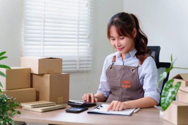 Merkez ofiste çalışan Asyalı genç kadın girişimci, çevrimiçi iş e-ticaret ve nakliye teslimatının ürün fiyatını hesaplamak için bir hesap makinesi kullanıyor..