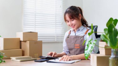 Önlüklü yetişkin bir Asyalı kadın online işi için finans hesapları yapıyor. Müşterileri için ev ofisinde nakliye ürünleri hazırlıyor..