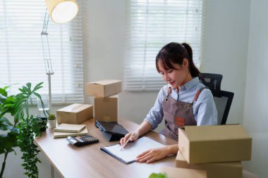 Genç Asyalı kadın, evindeki küçük işletmesinde çalışıyor. Evrak işlemlerini tabletle kontrol ediyor. Paket kutuları nakliyeye hazır..