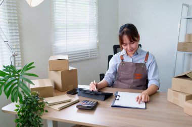 Önlüklü genç Asyalı kadın siparişleri internetten alıyor ve e-ticaret maliyetlerini hesaplamak için tablet kullanıyor. Nakliye ve teslimat için kutuları hazırlıyor..