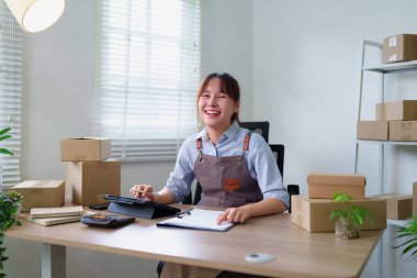 Genç Asyalı kadın siparişleri ve paketlemeyi hazırlayıp internet işini yönetiyor. Tablet ve belgelerini incelerken gülümsüyor..