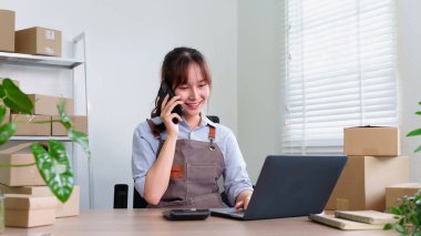 Bu ev ofisinden çalışan Asyalı genç bir girişimcinin fotoğrafı. Bilgisayarından siparişleri yönetirken telefonundaki müşteriyle gülümsüyor..