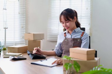 Evde tablet üzerinde çalışan Asyalı genç bir kadın. Eşyaları kutulara koyup müşterilere göndermeye hazır. Küçük işiyle eğleniyor..