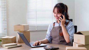 Asyalı genç girişimci müşterileriyle telefonda konuşarak online siparişleri onaylıyor. Bilgisayarını online iş yapmak ve siparişleri yönetmek için kullanıyor.