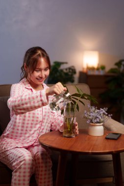 Asyalı yetişkin bir kadın, sakince ve düşünceli bir yaşam tarzını geliştirmek için içinde bitki yetiştirilen cam sürahisinden bir vazoya temiz su döküyor..