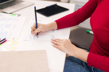 Asyalı yetişkin bir kadının eli kağıda kullanıcı arayüzü çiziyor. İş yerindeki beyaz bir masada mobil uygulama geliştirme için kusursuz bir kullanıcı deneyimi kavramsallaştırmak.