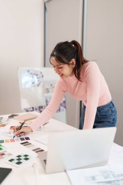 Genç bir Asyalı kadın yaratıcı stüdyosunda UI UX tasarımcısı olarak çalışıyor. Web sitesi ve mobil uygulama tasarımı için ilham almak için bir dizüstü bilgisayar ve renk paleti kullanır.