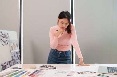 Genç Asyalı kadın UI tasarımcısı bir renk paleti, dizüstü bilgisayar ve çizimler kullanarak düşünceli bir proje planlıyor. Ofisteki renk eşleşmesi için kullanıcı arayüzleri tasarlanıyor