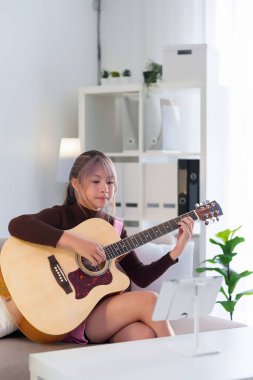 Genç bir Asyalı kadın kanepede oturur, gitar çalar ve tabletle internetten ders çalışır. Akustik gitar çalmayı, rahat evinde vakit geçirmeyi seviyor..