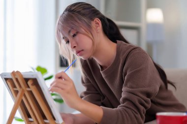 Yetişkin bir Asyalı kadın evde resim dersinde boya fırçasıyla tuvale resim yaparken görülüyor. O, sanat için yaratıcı hobi becerileri keşfetmek için kapalı alanda resim yapıyor.