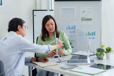 Asyalı iş adamları grafikleri analiz eder ve sürdürülebilir gelişimi temsil eden bir rüzgar türbini modelini tartışır. Çevresel sosyal yönetim girişimleri yeşil çözümler kurumsal sorumluluklar