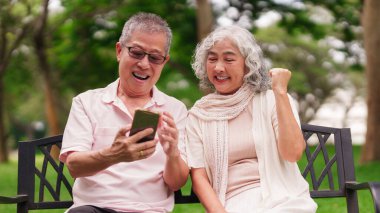 Parktaki bankta oturan mutlu Asyalı yaşlı çift akıllı telefon ekranında emekliliğin tadını çıkarın.