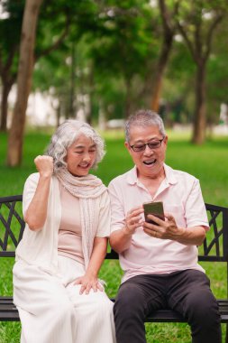 Neşeli yaşlı Asyalı çift parkta bankta oturmuş doğayla çevrili cep telefonlarını izliyorlar.