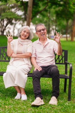 Gülümseyen Asyalı yaşlı çift parktaki bankta dinleniyor. Onaylanmış bir jest gösteriyorlar. Mutlu bir emeklilik hayatı.