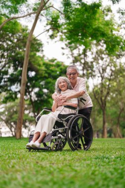 Mutlu Asyalı çift parkta güneşli bir günün tadını çıkarıyor Tekerlekli sandalye kullanıcısı eş emekliliğinden sevgi dolu destek alıyor dışarıda doğa