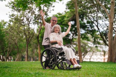 Yaşlı Asyalı koca, güzel yeşil park ortamında güneşin tadını çıkaran tekerlekli sandalyedeki karısına değer veriyor.