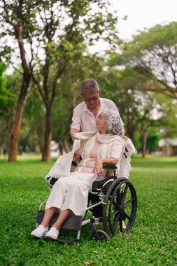 Asyalı koca, yeşil parktaki dinlenme gününde eşarbı tekerlekli sandalyeyle örtmeye yardım ediyor.