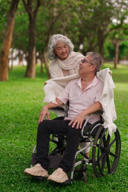 Asyalı yaşlı bir çift parkta sevgi dolu bir anı paylaşıyor. Tekerlekli sandalye, bakımı sembolize ediyor ve emeklilik yoldaşlığını devam ettiriyor.