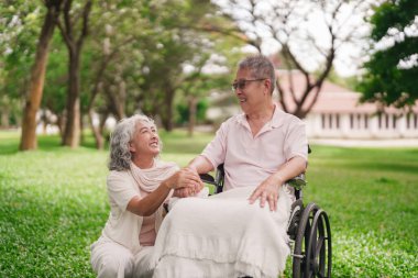 Asyalı yaşlı çift, mutlu an parkı kadını, el ele tutuşan koca tekerlekli sandalyesini destekliyor.