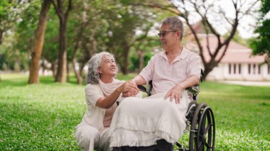 Asyalı emekli kadın tekerlekli sandalyede kocasının yanında diz çöker. El ele tutuşur. Sevgisini gösterir.