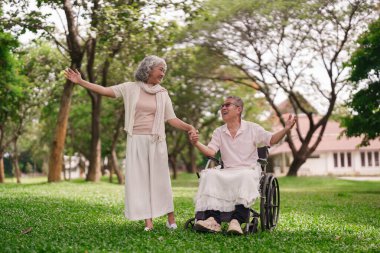 Mutlu Asyalı yaşlı çift parkta eğleniyor. Karısı, tekerlekli sandalyedeki kocasını destekliyor. Güzel bir aşk fotoğrafı, pozitif yaşlanma.
