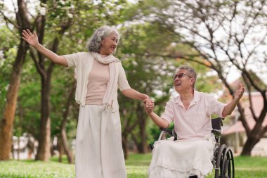 Açık hava parkında birlikte gülümseyen Asyalı son sınıf çifti. Tekerlekli sandalyedeki yaşlı adam bir kadınla el ele tutuşup doğanın tadını çıkarıyor..
