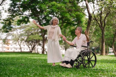Yaşlı Asyalı çift park zamanını sever. Tekerlekli sandalyedeki adam karısının elini tutuyor. Mutlu emeklilik anları, doğa sevgisiyle..