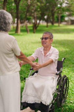 Yaşlı Asyalı çift, barışçıl parkta el ele tutuştukları özel anı paylaşıyor. Tekerlekli sandalyede oturan adam, karısından sevgi desteği alıyor. Emeklilik süresince bağlılık gösteriyor.